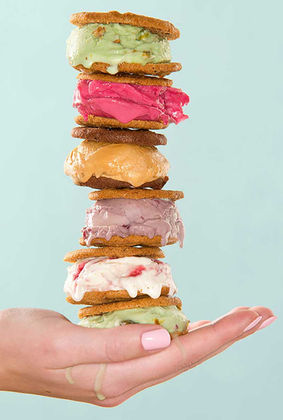 A hand holds a precariously stacked tower of ice cream sandwiches, featuring scoops of vibrant green, pink, caramel, purple, and white ice cream between golden-brown cookies. A small drip of melted ice cream is visible on the person's hand, set against a pastel blue background.