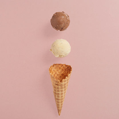 A deconstructed ice cream cone featuring a waffle cone at the bottom with two floating scoops of ice cream—one chocolate and one vanilla—against a soft pink background.