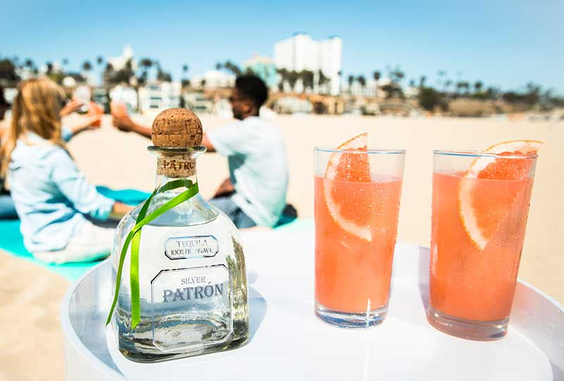 Two pink cocktails with a bottle of Patrón tequila on a sunny beach with people relaxing in the background