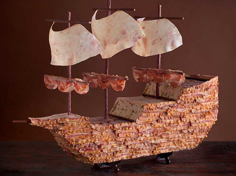 A detailed ship sculpture crafted entirely from cured meats, featuring a hull made of layered bacon, masts of sausage, and sails of thinly sliced deli meat, set against a dark brown background.