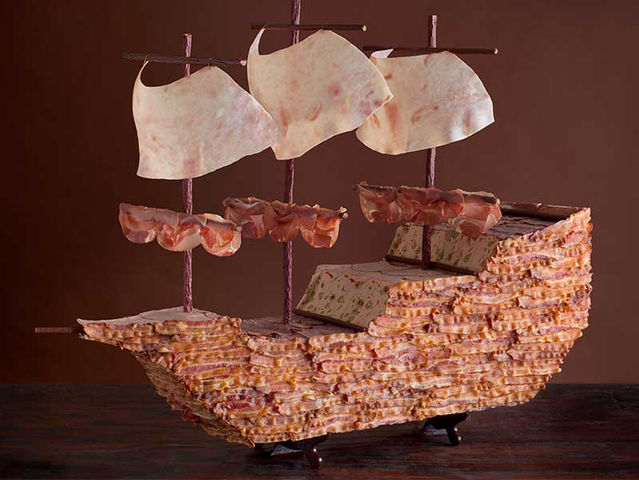 A detailed ship sculpture crafted entirely from cured meats, featuring a hull made of layered bacon, masts of sausage, and sails of thinly sliced deli meat, set against a dark brown background.
