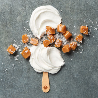 A creative composition featuring a wooden popsicle stick topped with swirls of white cream, scattered caramel cubes, and flakes of sea salt on a textured gray surface.