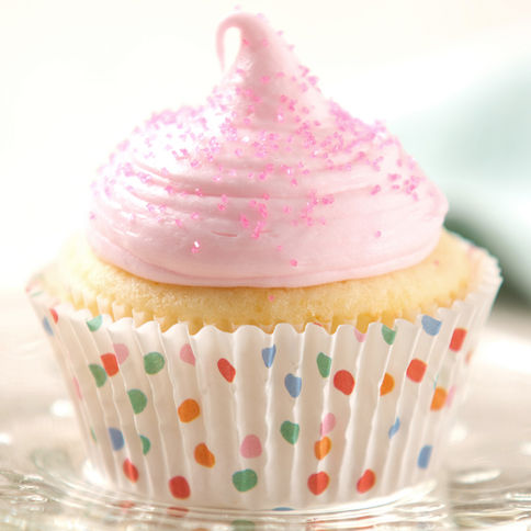 A fluffy vanilla cupcake in a colorful polka dot wrapper, topped with a swirl of pink frosting and a sprinkle of pink sugar, set on a glass plate.