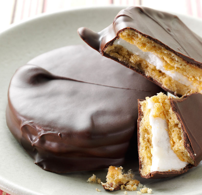 A plate with thick, round chocolate-covered cookies, one of which is broken open to reveal a layer of fluffy marshmallow and crispy graham cracker inside.