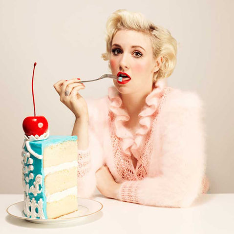 A woman with platinum blonde curls, wearing a soft pink fuzzy sweater, poses with a fork near her lips in front of a towering slice of blue-frosted cake topped with a glossy cherry.