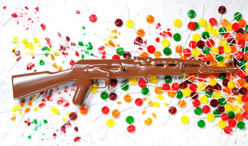 A life-sized rifle made entirely of glossy chocolate is placed on a marble surface, surrounded by shattered colorful lollipops in red, yellow, and green.