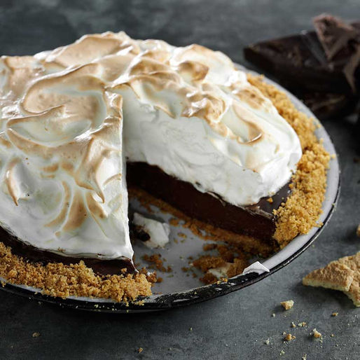 A rich chocolate pie with a graham cracker crust, topped with toasted meringue, sits on a dark surface with a missing slice revealing its velvety filling. Crumbs and chocolate pieces surround the dish.