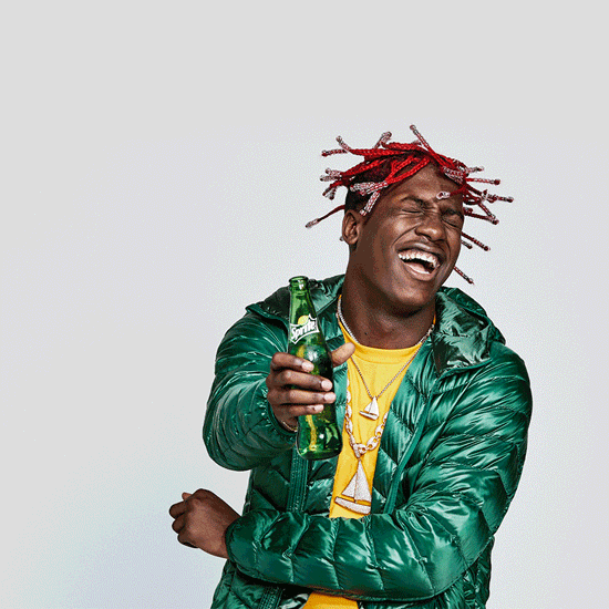 A young man with red braids and a big smile wears a green puffer jacket over a yellow shirt, holding a green glass bottle while laughing against a light gray background.