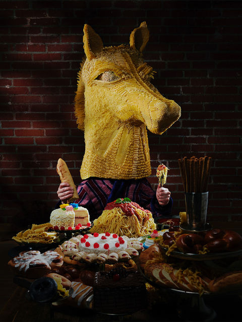 A figure wearing a large, pasta horse head sits at a dimly lit table overflowing with pasta, bread, cakes, and desserts, holding a baguette in one hand and a fork in the other, against a brick wall backdrop.