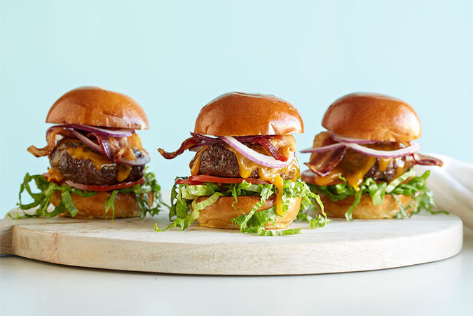 Three mini bacon cheeseburgers with lettuce, tomato, cheddar, red onion, and brioche buns arranged on a wooden board.