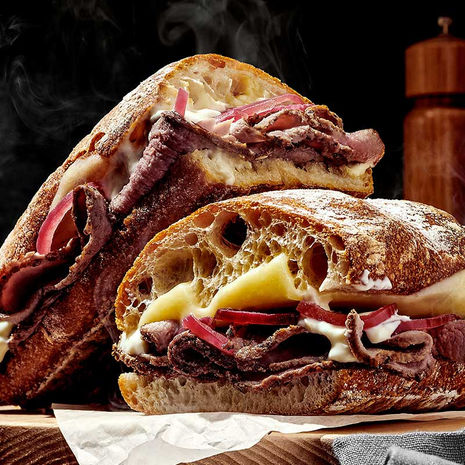 A crusty artisan sandwich filled with tender roast beef, melted cheese, and pickled red onions, cut in half and stacked, with steam rising from the warm bread.