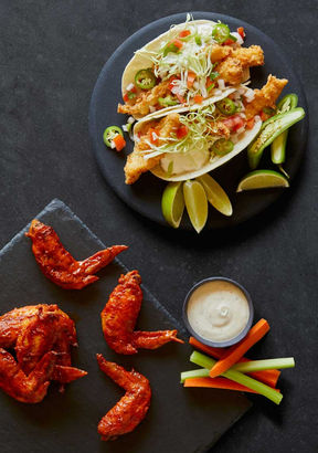A plate of crispy fish tacos topped with shredded lettuce, diced tomatoes, jalapeños, and lime wedges sits next to a slate board with buffalo wings, celery and carrot sticks, and a cup of creamy dipping sauce.
