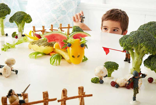 A playful food art scene featuring a dragon made from yellow bell peppers, watermelon, and leafy greens, surrounded by broccoli trees, mushroom soldiers, and tiny toy knights engaged in battle, while a young boy watches from behind a barrier.