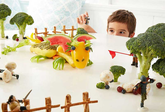 A playful food art scene featuring a dragon made from yellow bell peppers, watermelon, and leafy greens, surrounded by broccoli trees, mushroom soldiers, and tiny toy knights engaged in battle, while a young boy watches from behind a barrier.