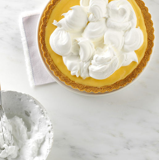 A golden graham cracker crust holds a smooth lemon filling, topped with dollops of freshly whipped cream. A bowl with remnants of whipped cream sits nearby on a marble countertop.