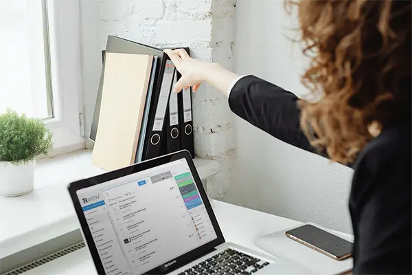 Laptop zeigt Archivierungssoftware SF Archiv. Vor dem Laptop sitzt eine Frau. Diese greift aber noch zu den Akten auf der Fensterbank – arbeitet also noch nicht komplett digital