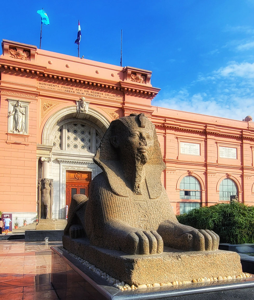A sphinx statue sits outside a pink historic building under a clear blue sky, with flags and detailed carvings visible.