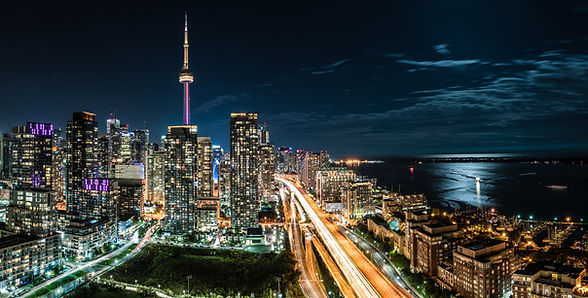 Toronto city skyline at night, with the CN tower illuminated pink and the glow of cars rushing along the Gardiner expressway. The moon illuminates Lake Ontario