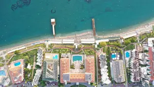 Аэрофотоснимок курортного комплекса Fame Residence Kemer & SPA 5* в Турции. Отель с несколькими бассейнами и зеленой территорией расположен непосредственно на берегу Средиземного моря. Видны пляжная зона с пирсами, основное здание с терракотовой крышей и прилегающие корпуса.