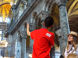 Группа туристов слушает экскурсовода в красной форме Saodat Turizm внутри Айя-Софии в Стамбуле. Он указывает на архитектурные детали, а туристы внимательно слушают, не отвлекаясь на телефоны. Внутреннее убранство включает колонны, арки, мозаики и исламскую каллиграфию, освещённые большими люстрами.