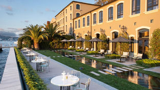 Террасы отеля Vakko Hotel Sumahan Bosphorus на берегу Босфора с декоративными водными каналами, зелеными газонами, белыми обеденными столиками и зонтами, на фоне исторического здания османской винокурни золотистого цвета и пальм