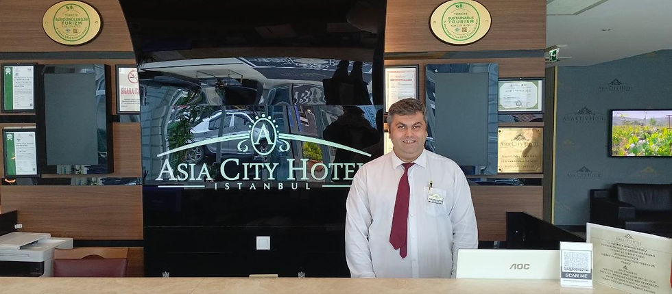A man in a white shirt and burgundy tie is standing at the reception desk of the Asia City Hotel Istanbul. Certificates hang on the wall.