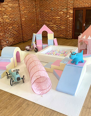 Pastel coloured soft play set up indoors. Includes pastel pink, blue and yellow foam shapes, large white square ballpit, pink pop up tent and tunnel, blue ride on vintage car, pink ride on scooter and blue bouncy hopper on white foam matting.