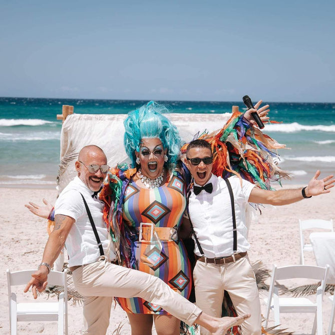 Vibrant drag queen with stunning blue hair celebrating at a beach wedding with two joyful grooms.