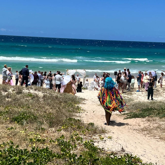 Resplendent drag queen in a vibrant rainbow outfit, strolling down the beach towards a joyous same-sex wedding, with all guests in attendance.