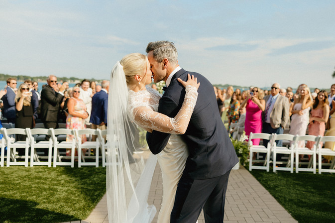 bride and groom kiss at the end of the aisle with guests clapping and cheering in the background
