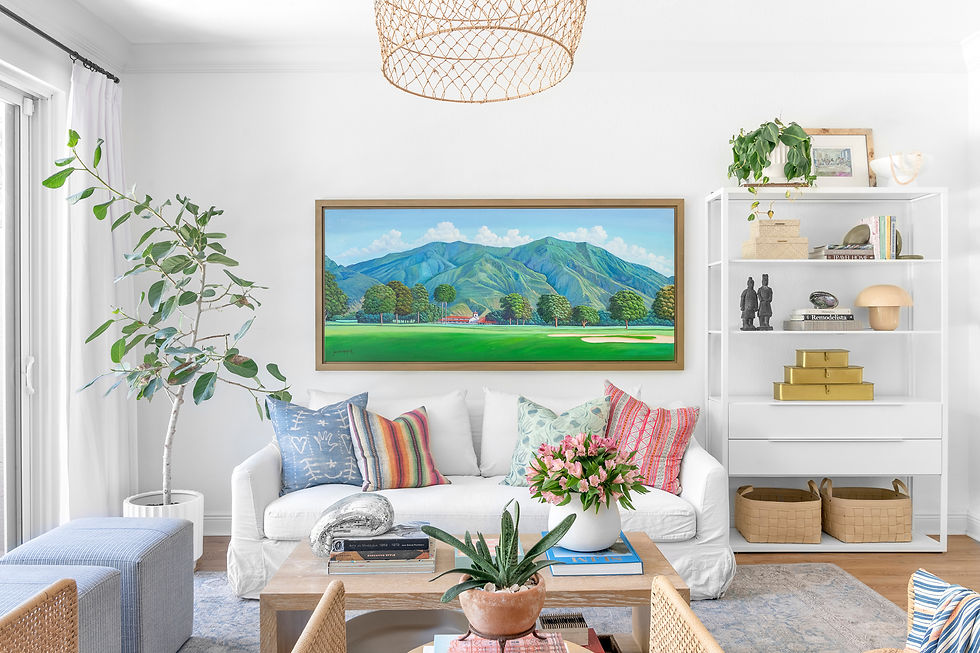 Bright living room with a white sofa, colorful pillows, and plants. A painting of mountains hangs on the wall. White shelves hold decor.