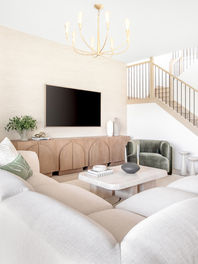 Modern living room with beige sofa, green armchair, TV on wood panel wall, and staircase. Light decor, plants, and gold chandelier by Studio 790.