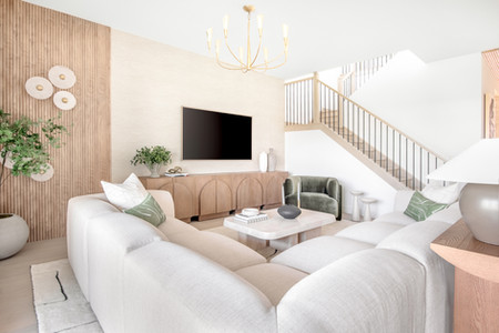 Modern living room with cream sofas, green chair, large TV, and wooden accents. Sunlit space with plants and decorative vases. Cozy vibe.