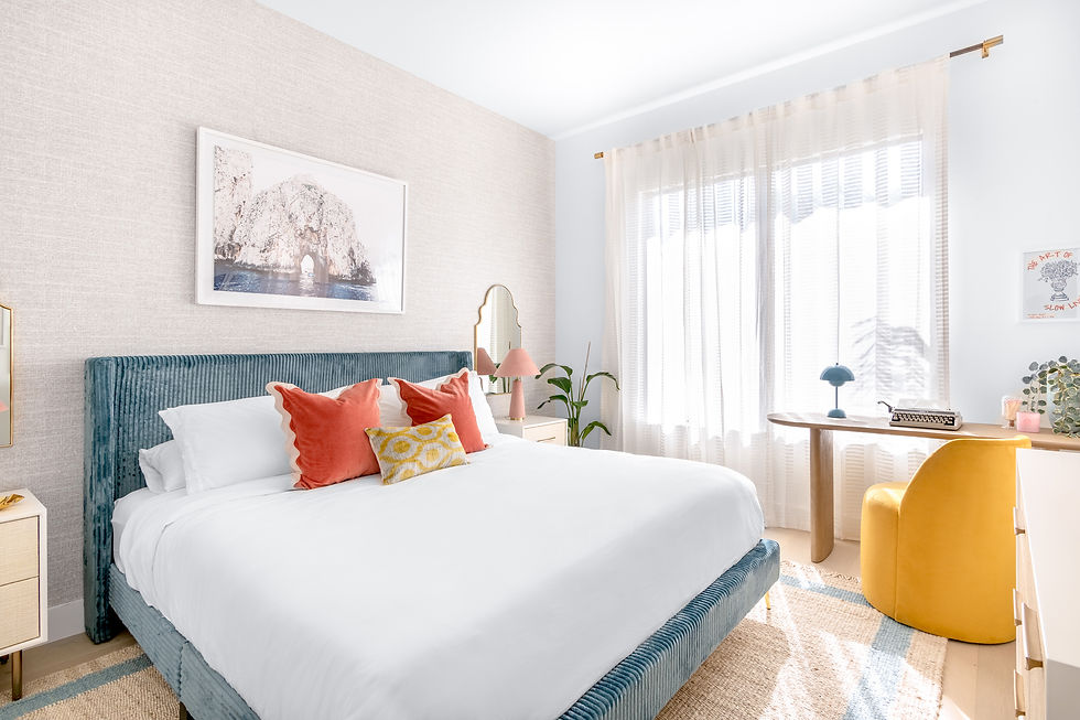 Bright bedroom with teal bed, white linens, orange pillows, and art. Sunlit curtains, wood desk with typewriter, yellow chair. Cozy vibe.