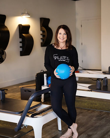 Pilates on the Plains owner, Becky Anders, stands next to Pilates reformers holding a blue exercise ball. 