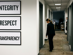 An abstract conceptual image showing a beautifully framed corporate "Values" poster hanging on a wall, contrasting with a long, dimly lit office hallway stretching into the background, symbolizing the gap between stated company values and actual workplace culture.