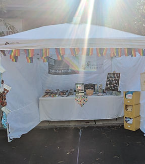 Tent and Table vendor setup at the Mt. Airy Village Fair. Behind the table is a banner with information to find this website. On the table are a variety of handmade sensory and home goods, created by Ev and their partner, Jeremy.