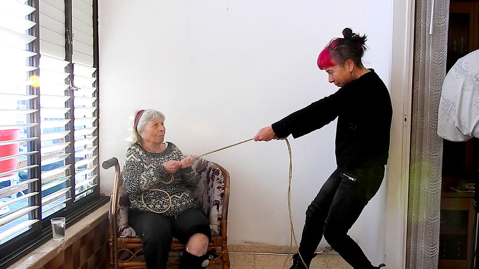 צילום של אשה עומדת שמושכת חבל אותו מחזיקה אישה מבוגרת שיושבת על כורסה. לצד האישה המבוגרת מקל הליכה.