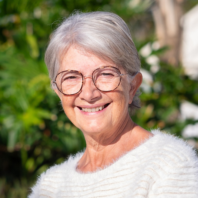 Femme âgée souriante aux lunettes et cheveux gris, en extérieur ensoleillé.