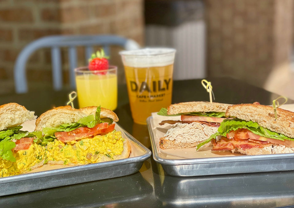 two gourmet sandwiches on silver trays next to an orange juice and a beer in a Daily branded cup