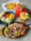 Fajitas with lime on a skillet, a plate with tortillas, cheese, and veggies, chips, red and yellow drinks with lime, on a gray table.