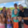 A group of smiling people on a boat, with a child holding a crab. Green marshland and blue sky in the background. Casual, joyful mood.