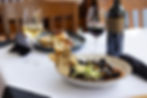 Restaurant table with bowl of mussels, bread, and wine glasses beside a bottle on a white tablecloth.
