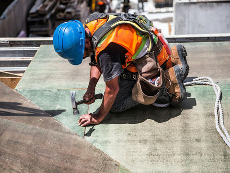 Construction worker in a blue hard hat, orange safety vest, and harness kneels on a jobsite surface while hammering material into place, illustrating the hands-on skilled labor behind Washington, D.C. construction projects.