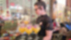 A woman in a lace top serves Mama Mango drinks garnished with purple flowers on a tray. The festive background features string lights and colorful decor.