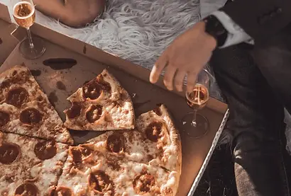 Pizza on a fluffy carpet, a man and woman sitting next to it with glasses of champagne. Spicy Fake Dating Romance.