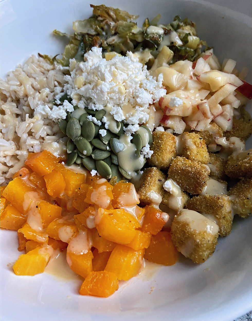 A grain bowl with brown rice, roasted butternut squash, crispy tofu, chopped apple, roasted Brussels sprouts, pepitas, feta, and a maple Dijon dressing.
