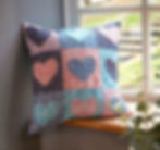 Patchwork cushion on windowsill, featuring heart patterns in blue, pink, and floral prints. Sunlight filters through the window, creating a cozy mood.