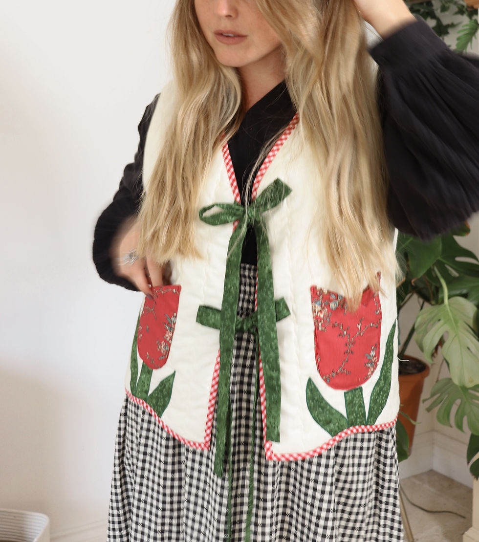 A person wearing a white vest with red tulip pockets and green ties over a black top. Background features a green plant, creating a cozy vibe.