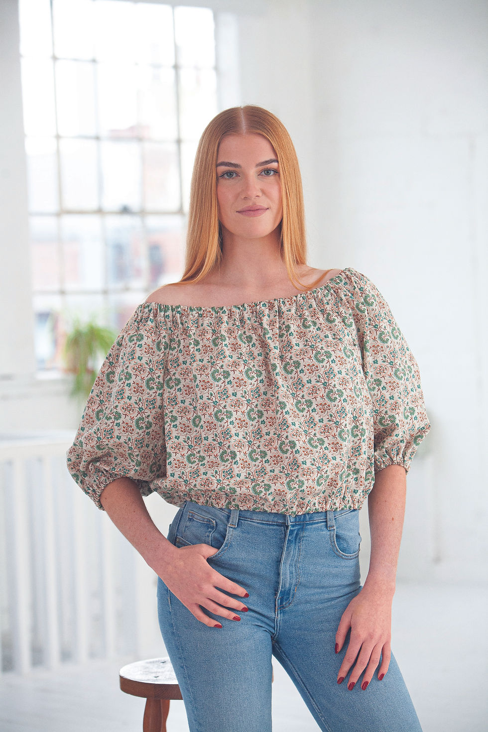 Woman in a floral off-shoulder blouse and blue jeans poses in a bright room, hand on hip, with a soft smile.